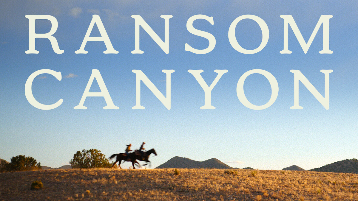 Duas pessoas montadas a cavalo atravessam uma colina coberta de gramíneas sob um céu azul-claro. As palavras ‘Ransom Canyon’ se destacam em letras grandes que ocupam a parte superior da imagem.