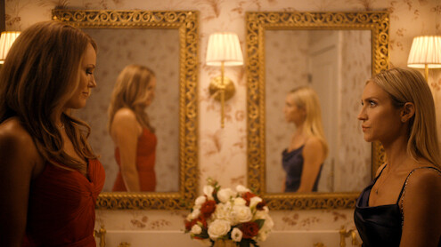 Two women in elegant dresses face each other in a decorative, warmly lit bathroom with floral wallpaper, ornate mirrors, wall lamps, and a bouquet of flowers on the sink between them. Their reflections appear in the mirrors.