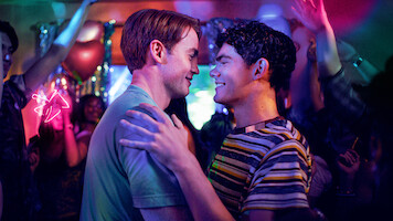 Two men smiling and dancing closely together at a vibrant, colorful party with people celebrating, arms raised, and festive decorations in the background under neon and stage lights.