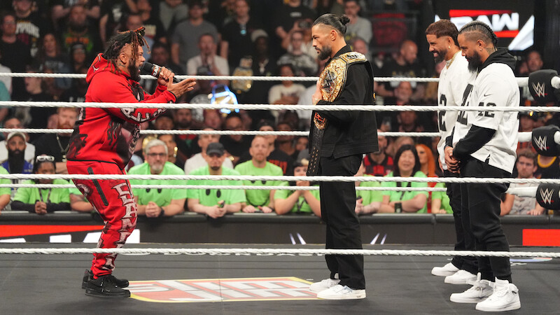 Four wrestlers face off in a WWE ring with a large audience in the background, one in red holding a mic, one wearing a championship belt, and two in matching outfits, during a dramatic segment of a wrestling event.