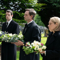 Three solemn people in black coats holding flower arrangements stand in a cemetery, surrounded by gravestones and green trees, attending a funeral on a cloudy day.