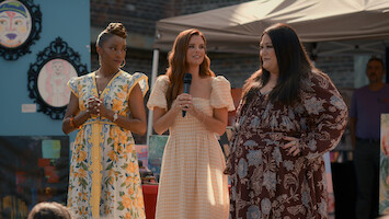 Three women in summer dresses stand together outdoors at an art fair or festival, one holding a microphone. Colorful paintings and a canopy tent are visible in the background.