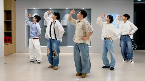 Cinco pessoas com roupas casuais posam em um estúdio de dança com piso de madeira clara, decoração moderna e um letreiro de néon na parede, todas fazendo a mesma pose da coreografia e voltadas para a esquerda.