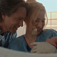 A smiling man and woman in medical scrubs hold a newborn baby together in a brightly lit room, expressing joy and tenderness after childbirth.