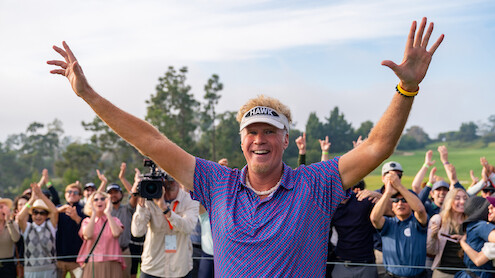 Smiling golfer with raised arms celebrates on a golf course, surrounded by cheering spectators and trees in the background, under a partly cloudy sky.