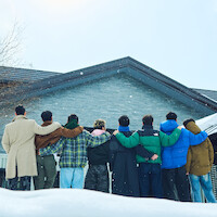 A group of eight people stand arm-in-arm, facing a snow-covered house, dressed warmly in winter clothing. The environment is wintry and serene, conveying friendship and togetherness in a peaceful outdoor setting.