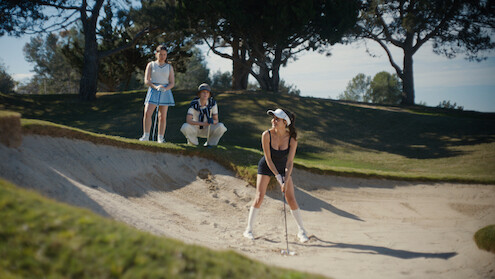 Mikaela Hoover as Ava prepares to hit a golf ball from a sand bunker as two people watch nearby on a sunny golf course surrounded by trees and grass.
