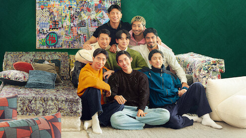 The cast of 'The Boyfriend' Season 2 sit closely together on a colorful sofa in a cozy living room with eclectic pillows, patterned furniture, and a vibrant art piece on a green wall, smiling and posing for the photo.