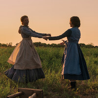 Duas mulheres em vestidos antigos dão as mãos e giram alegremente em um gramado no pôr do sol, com a luz quente do sol deixando um brilho dourado sobre a paisagem rural.