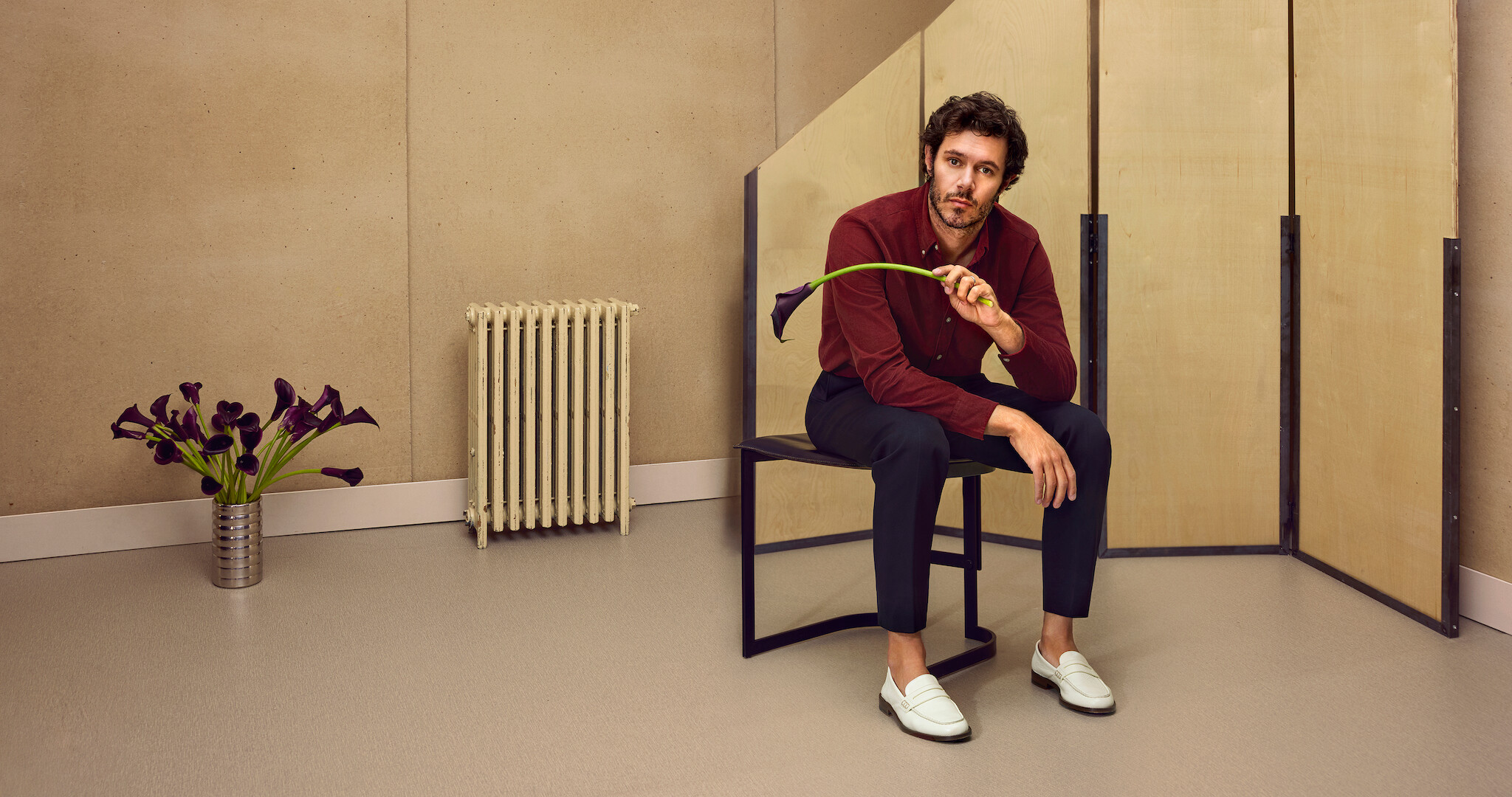 Adam Brody sits on a stool holding a black calla lily 