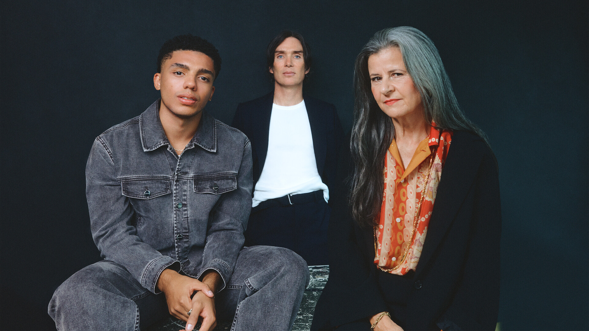 From left to right, Jay Lycurgo, Cillian Murphy, and Tracey Ullman sit around a table against a dark slate grey background.