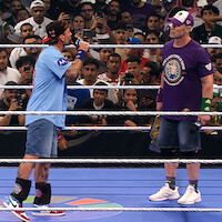 Two wrestlers face off in a crowded wrestling ring, one wearing a blue shirt speaking into a microphone, the other in a purple shirt holding a championship belt. The audience watches intently from all sides.