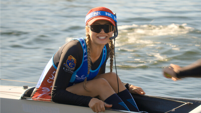 Smiling woman with braids wearing sunglasses, a visor, and athletic gear sits in a rowing boat on the water, holding equipment with one hand and a headset microphone, sunlight reflecting off the rippling water in the background.