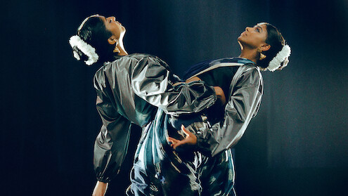 Two dancers in matching shiny costumes perform on stage with dramatic lighting, facing away from each other and leaning back-to-back, wearing white floral hair accessories in a dark, theatrical setting.