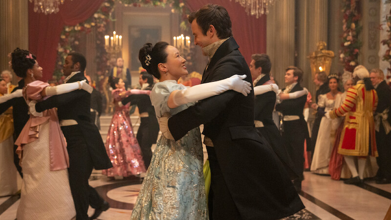 Um casal em elegantes trajes de época dança em um grande salão, cercado por outros casais dançando e convidados em vestes elaboradas, com decorações deslumbrantes, flores e lustres, em uma atmosfera festiva de época.
