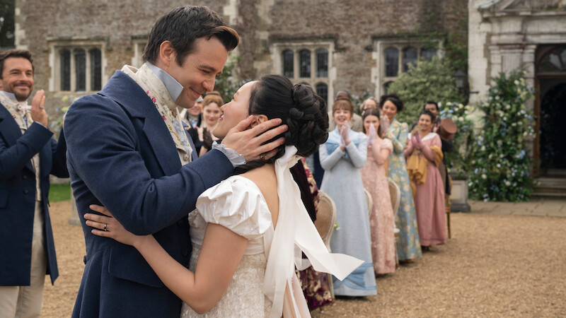 A bride and groom in period costumes embrace and smile outdoors, surrounded by cheering guests in Regency-era attire, with an old stone building and greenery in the background.