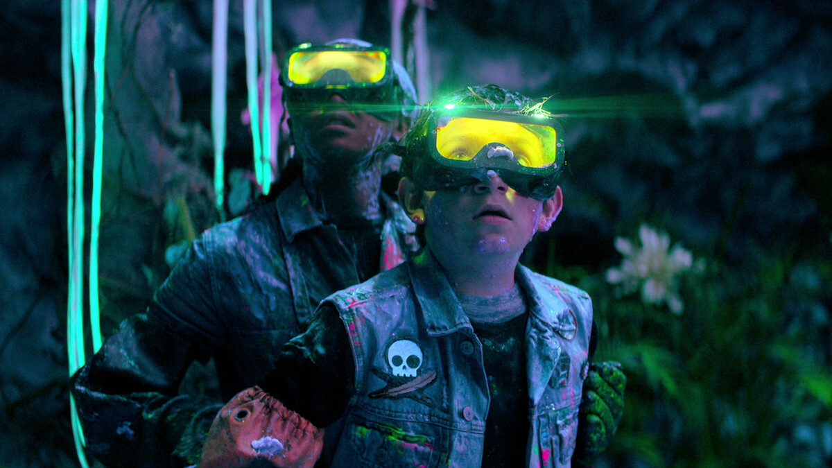 Two people wearing yellow glowing goggles and denim jackets with patches stand in a colorful, neon-lit cave environment, surrounded by plants and glowing decorations, looking upward as if intrigued or cautious.