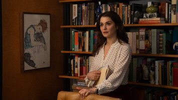 Rachel Weisz as The Protagonist  in a white blouse stands in front of a bookshelf filled with books and decor, holding folders. A framed artwork hangs on the wall, creating a cozy, intellectual office or library setting.