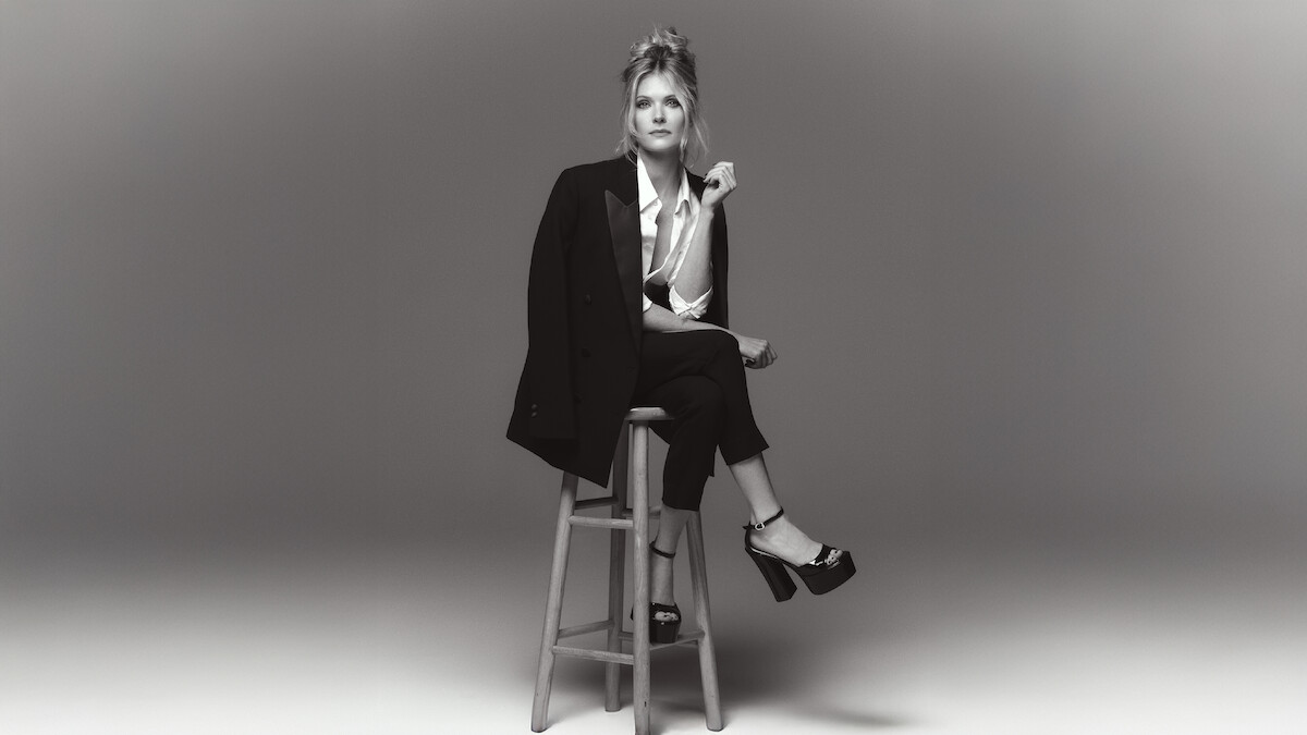 Meghann Fahy sits on a stool in a chic black suit with white button up blouse with legs crossed and platform heels.