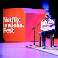 David Spade seated on stage with a microphone, speaking at a Netflix is a Joke Fest event. Bright stage lighting, large red event sign in background, wooden floor and casual atmosphere.
