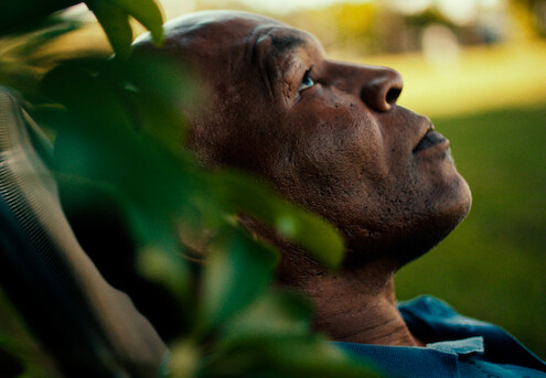 Close-up de um homem com uma tatuagem no rosto próximo a plantas em um jardim. Ele olha para cima, reflexivo