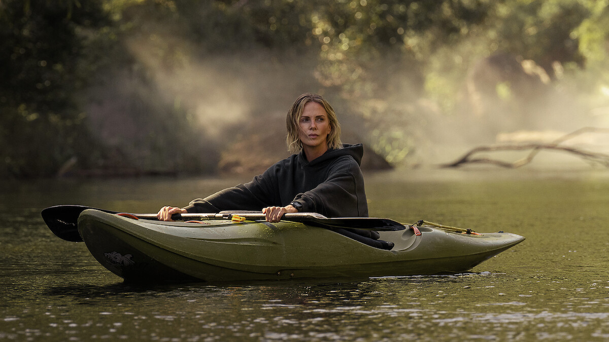 A person wearing a dark hoodie is paddling a green kayak on a calm river, surrounded by trees with mist rising in the background, creating a serene outdoor nature scene.