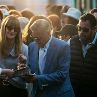 A man in a blue suit signs autographs in a crowded outdoor setting at sunset. People around him wear sunglasses and casual clothing, creating an excited and lively atmosphere.