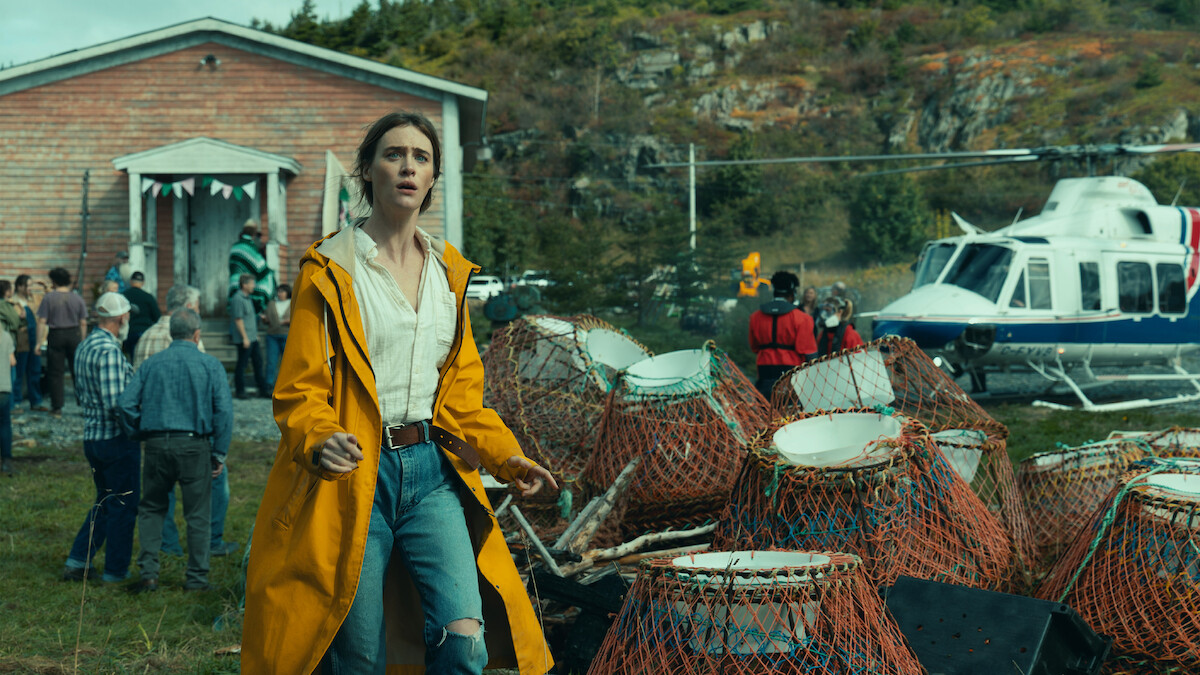 Woman in yellow raincoat stands near lobster traps, outdoors by a crowd and a helicopter, with a wooden building and forested rocky hill in the background, suggesting a coastal rural setting.