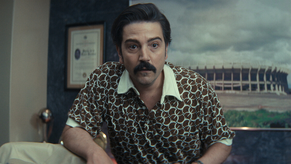 Man with dark hair and mustache in patterned shirt leans forward in an office with framed certificate on wall and large stadium photo in the background.