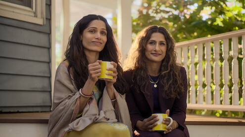 Two women sit together on a sunlit porch, holding yellow mugs and smiling. Shrubs and a white railing are visible in the background, creating a relaxed, inviting environment.