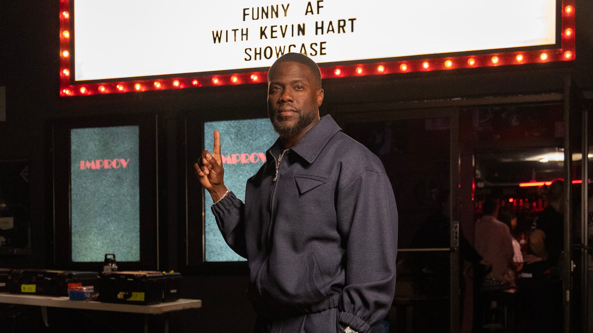 Kevin Hart in a blue jacket stands inside a comedy club, pointing upward at a lit marquee sign that reads “Funny AF with Kevin Hart Showcase,” with tables, equipment, and an audience in the background.