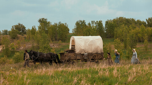 A covered wagon pulled by horses travels through a grassy field with three women and a child walking beside it, surrounded by trees under a partly cloudy sky.