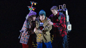 Three young people in winter clothes, holding ghost-busting equipment with glowing lights, stand closely together in a dark, dramatic setting, appearing focused and ready for action.