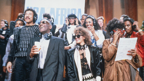 A diverse group of musicians in a recording studio, wearing headphones, singing and smiling together in front of a USA for Africa sign, with music sheets and microphones in a lively, collaborative atmosphere.