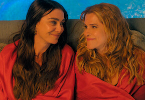 Duas mulheres estão sentadas em um sofá, envoltas em uma manta vermelha. Elas se entreolham, sorrindo