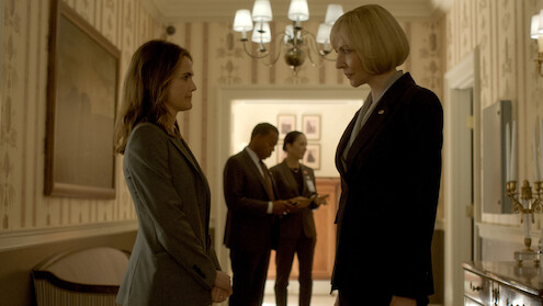 Two women in business suits face each other in a well-lit hallway with elegant decor; two other people are talking in the background, suggesting a formal or professional setting.
