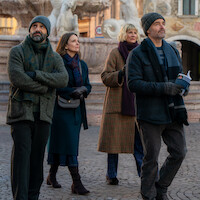 Four adults in winter clothing walk together in a cobblestone plaza, with an ornate fountain and historic buildings in the background, suggesting a city sightseeing setting in cold weather.