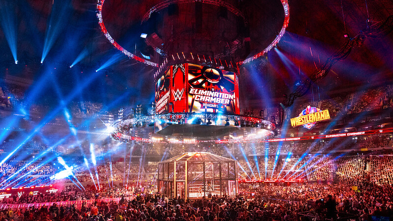 WWE Elimination Chamber wrestling event in a large arena, filled with a cheering crowd, colorful lights, a cage structure in the ring, and giant display screens with event logos and a WrestleMania sign in the background.