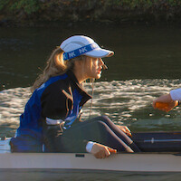 A female coxswain in a blue and black jacket and white cap sits in a rowing boat on the water, holding a microphone, with another rower’s hand visible, surrounded by natural outdoor scenery.