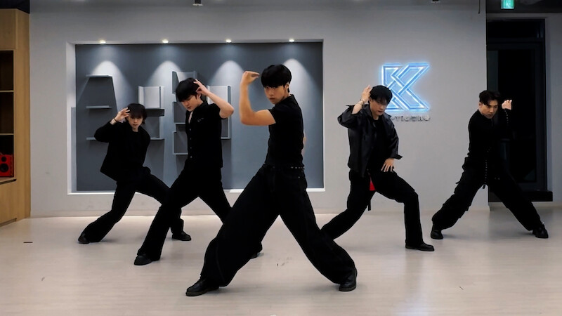 Five dancers in black outfits pose mid-routine in a modern dance studio with bright ceiling lights and a glowing K logo on the wall, light wooden floors, and geometric wall decor in the background.