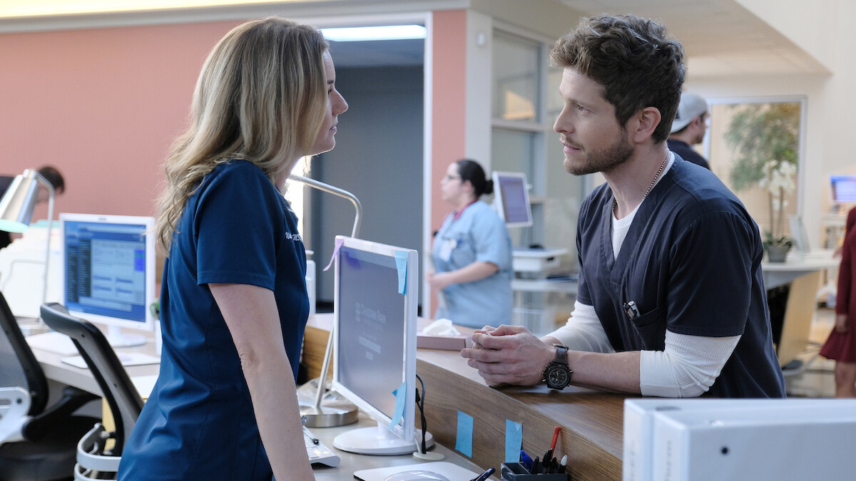 Two medical professionals in scrubs converse at a hospital reception desk with computers; other staff work in the background in a bright, modern clinical environment.