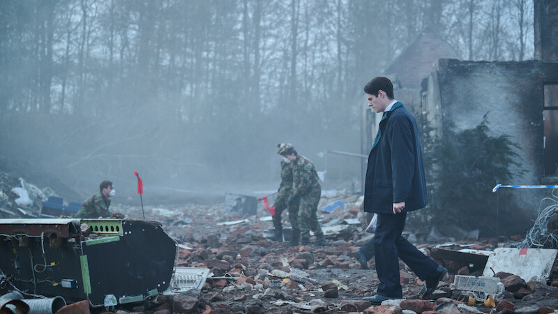 A man in a suit walks through a debris-strewn, foggy ruin as two people in camouflage uniforms investigate the wreckage; trees and a partially destroyed building are in the background.