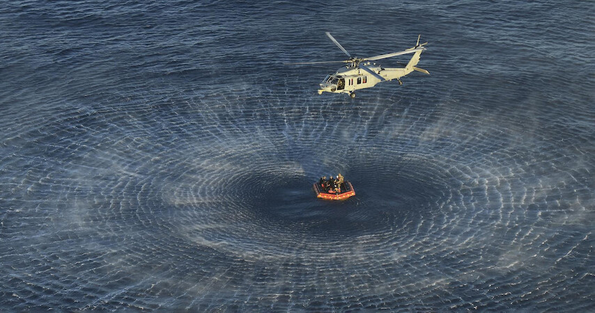 A helicopter hovers above the ocean, creating circular waves while performing a rescue operation near a life raft with several people aboard.