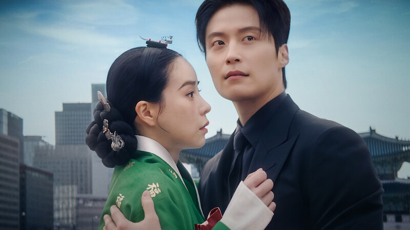 A woman in traditional Korean hanbok and a man in a modern black suit embrace with concern, standing outdoors against a backdrop of city skyscrapers and historical palace architecture under a blue sky.