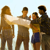 Four young people stand in tall grass outdoors, closely examining a large map together in bright daylight, appearing focused and collaborative in a natural setting.
