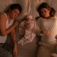 A man and woman lie on a bed with a newborn baby swaddled between them. The setting is a bedroom with warm lighting, suggesting a family moment. The woman holds a thermometer, possibly indicating concern for the baby's health.