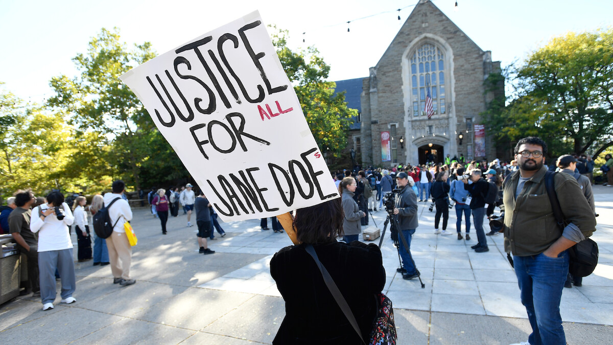 Person holds "Justice for Jane Doe" sign at outdoor protest in front of a large stone building, with a crowd of people gathered around under a sunny sky with trees in the background.