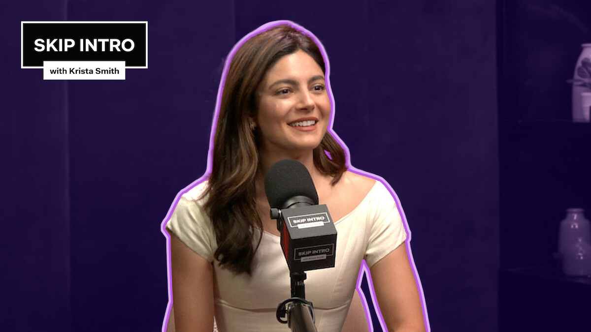 Monica Barbaro smiles in front of a microphone, framed by a deep purple background with a matching purple outline surrounding her.