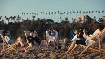 j-hope, Suga, Jin, RM, Jung Kook, and Jimin relaxing on beach chairs in the sand, enjoying a sunny day. Palm trees and buildings are visible in the background.