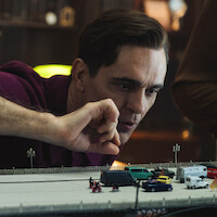 A man in a library or study leans over a detailed model of a bridge with miniature cars and people, carefully arranging the scene with his hand.